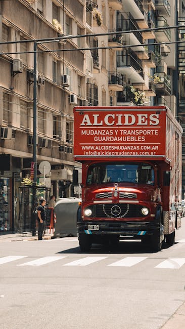 A street scene showing a small, clearly marked moving van for a house removal, operated by Movers Barnes, parked in a narrow urban road lined with multi-storey buildings with traditional architecture.