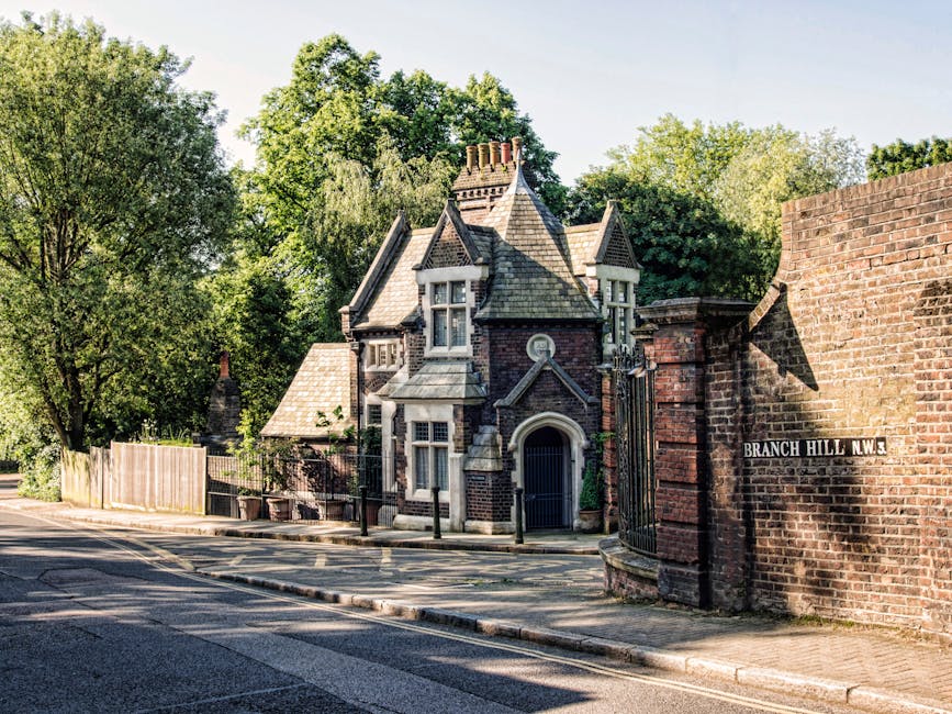 A narrow residential street view of a Victorian-style house situated behind a brick wall with a wrought iron gate, located on Church Road in Barnes. The house features ornate architectural details, including pointed gables, bay windows, and a steeply pitched roof covered in slate tiles. Surrounding the property are tall, leafy trees casting dappled shadows on the pavement and road, which has a double yellow line running along its edge. To the right, a street sign indicates 'Branch Hill N.W.3' attached to the brick wall. In the foreground, the street and pavement are visible, with the area appearing calm and quiet. The scene is illuminated by daylight, suggesting a clear, sunny day. This image is relevant for homeowners preparing for house removals, with potential logistical considerations for furniture transport and packing occurring within or around such Victorian homes, as managed by Movers Barnes.