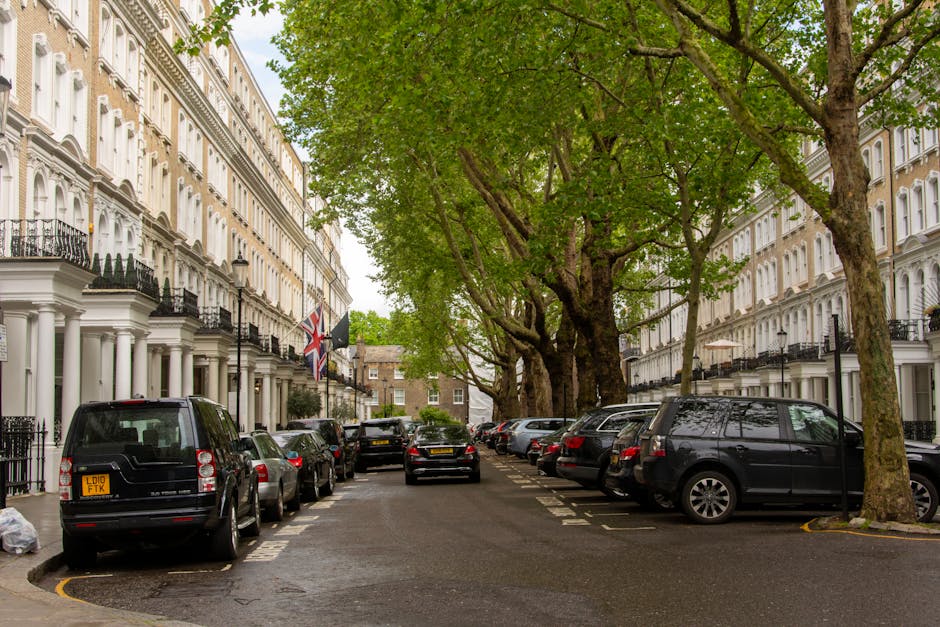 A residential street scene featuring white Victorian-style terraced houses with decorative moldings, black wrought iron railings, and balconies. The street is lined with large, leafy trees providing shade over parked vehicles, including black, silver, and grey cars parked along both sides of the narrow road. Some cars are parked close to the curb, while others extend slightly into the roadway. The pavement is wet, indicating recent rain, and a few plastic bags are visible on the sidewalk. In the background, a black van is positioned towards the center of the street, likely involved in house removals or furniture transport, as part of a home relocation process. The setting appears calm and organized, with domestic and moving logistics occurring in a typical urban residential area, aligning with the context of house removals and moving services offered by Movers Barnes, as described in the page about Victorian homes on Church Road Barnes.