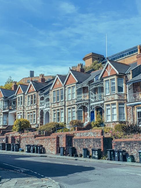 A row of traditional Victorian-style terraced houses on Lonsdale Road in Barnes SW13, featuring brick facades, ornate white-painted ironwork balconies, large bay windows, and gabled roofs. The houses are elevated above the street, with brick retaining walls and landscaped front gardens. On the pavement in front, several black and green refuse bins are lined up, and the street appears quiet with minimal traffic. The sky above is clear with some wispy clouds, and sunlight illuminates the residential façade, highlighting the architectural details. This scene is typical for area-specific house removals, with the architecture reflecting classic London residential design, suitable for furniture transport, packing and moving, and home relocation activities undertaken by professional removers such as Movers Barnes. The overall environment is neat and well-maintained, emphasizing the importance of careful logistics during house removals in the Barnes SW13 area.